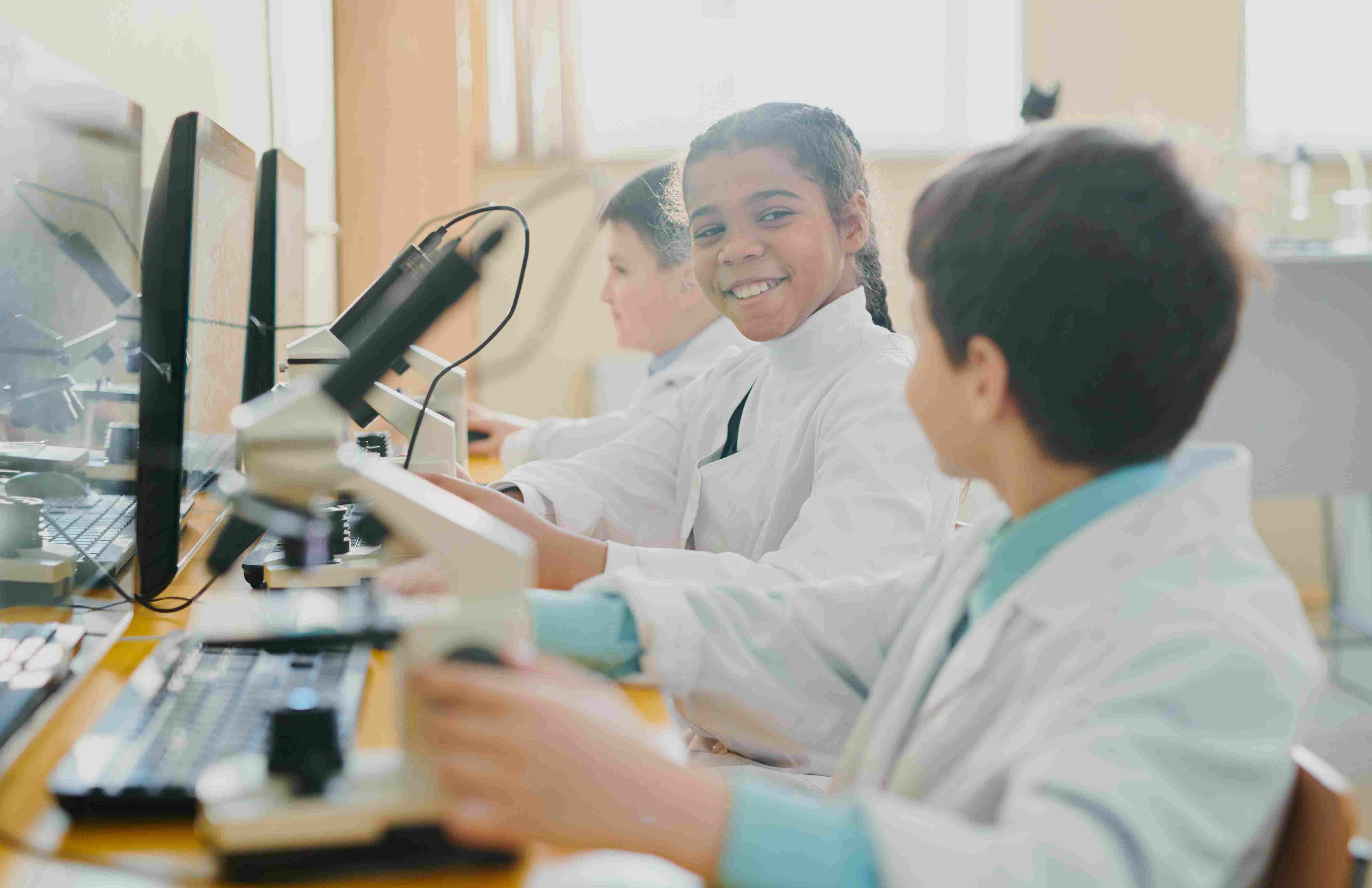 Drei Kinder, die in Laboruniform vor einem Computer und Mikroskop sitzen
